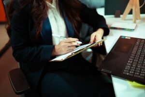 A focused businesswoman writing on a clipboard with a laptop nearby in an office environment.